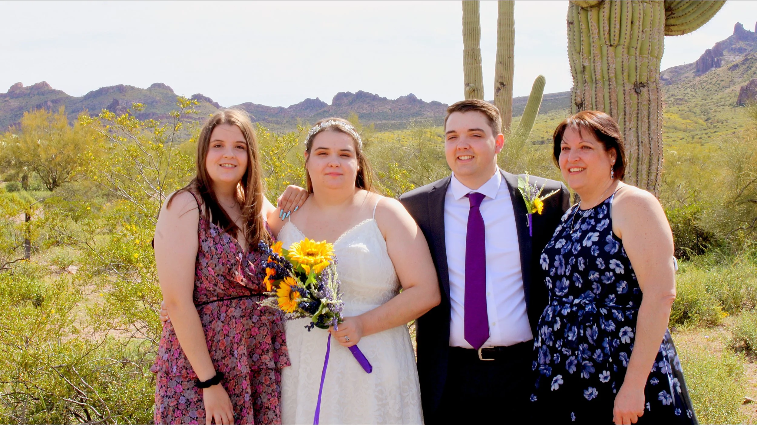 Group of five people standing outdoors in a desert landscape with mountains and cacti, smiling for a photo at a wedding or celebration.