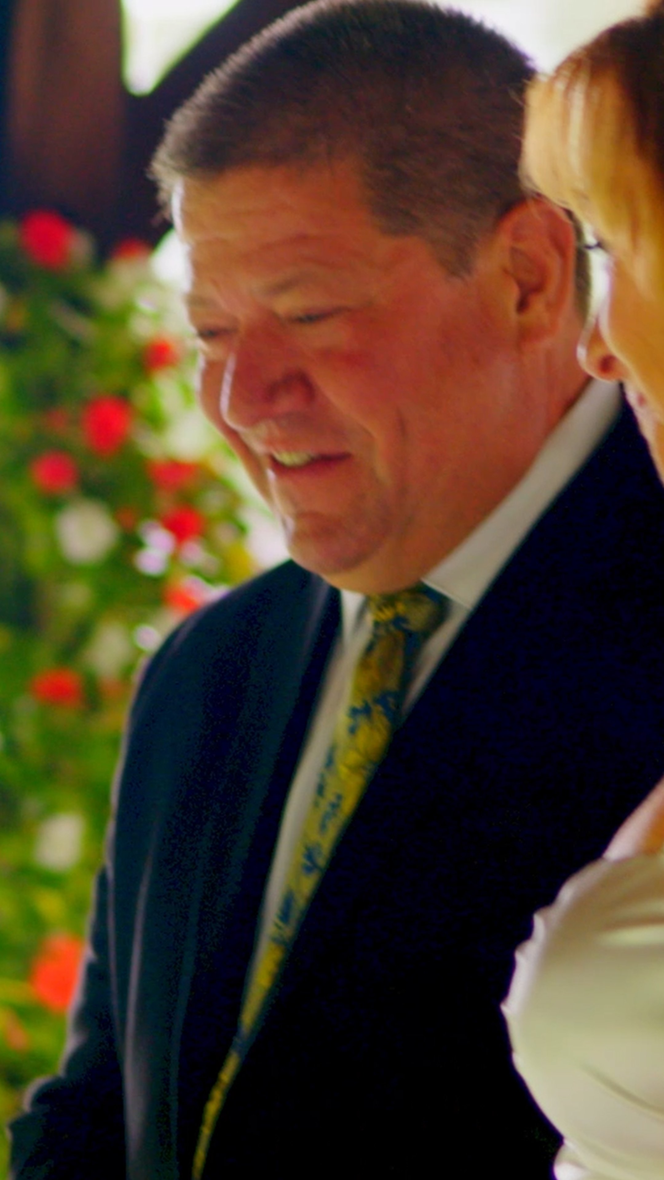 A man in a suit smiling, with a woman partially visible beside him, blurred flowers in the background.