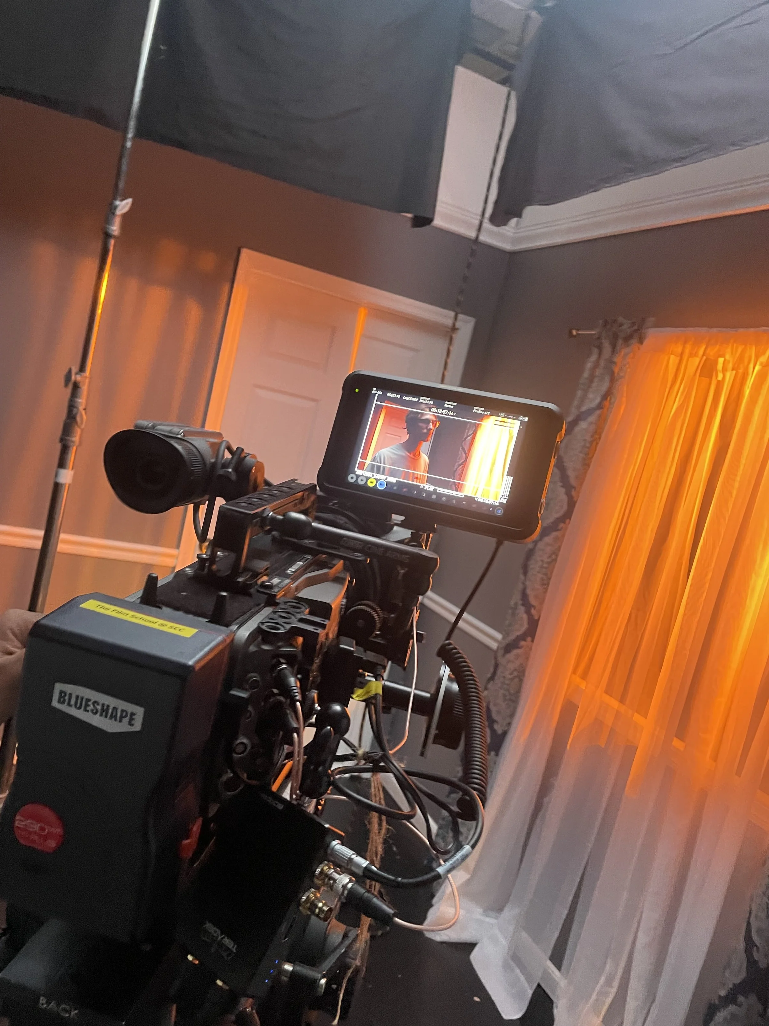 A film camera setup in a room with gray walls, orange curtains, and white trim. The camera's monitor shows a person wearing glasses and a light-colored shirt, standing near a window with yellow curtains.