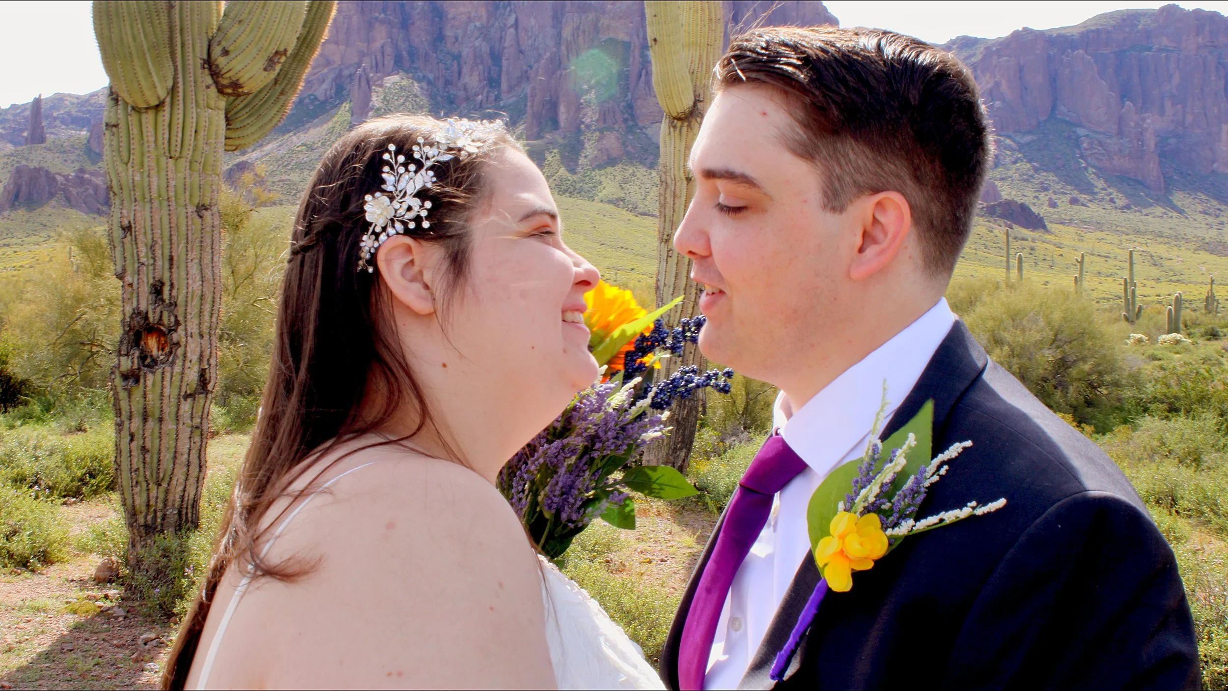 A bride and groom are close together outdoors in a desert setting with tall cacti, mountains, and a clear sky in the background. The bride has a floral headpiece and is smiling, while the groom is in a suit with a purple tie and floral boutonniere.