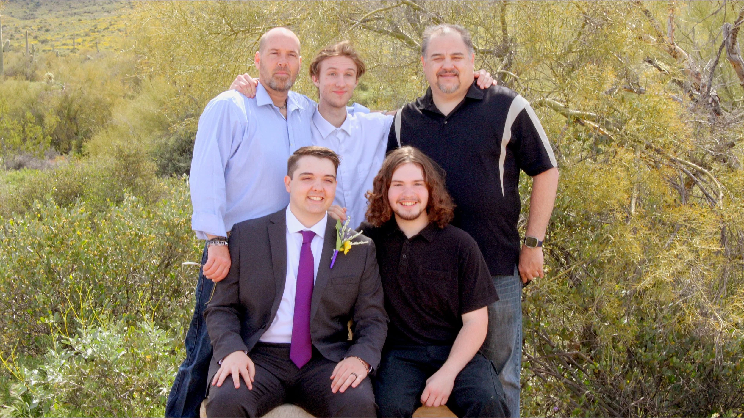 Six men are posing outdoors in a natural setting with trees and shrubs in the background, some with arms around each other.