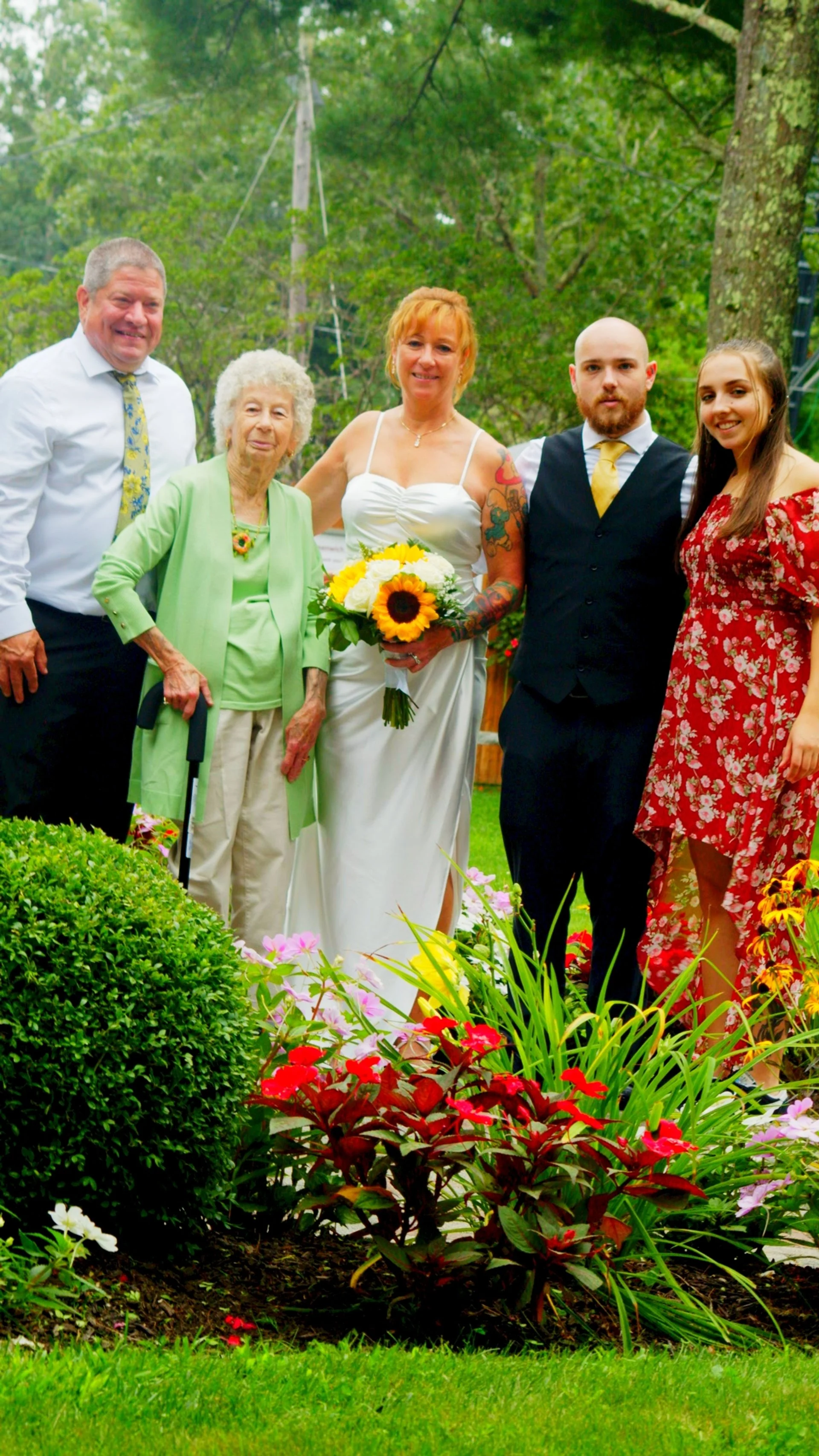 A group of six people, including an elderly woman with a cane, four adults, and one young woman in a red floral dress, standing outdoors in a garden with flowers and trees, celebrating a wedding with the bride in a white dress holding a sunflower bou