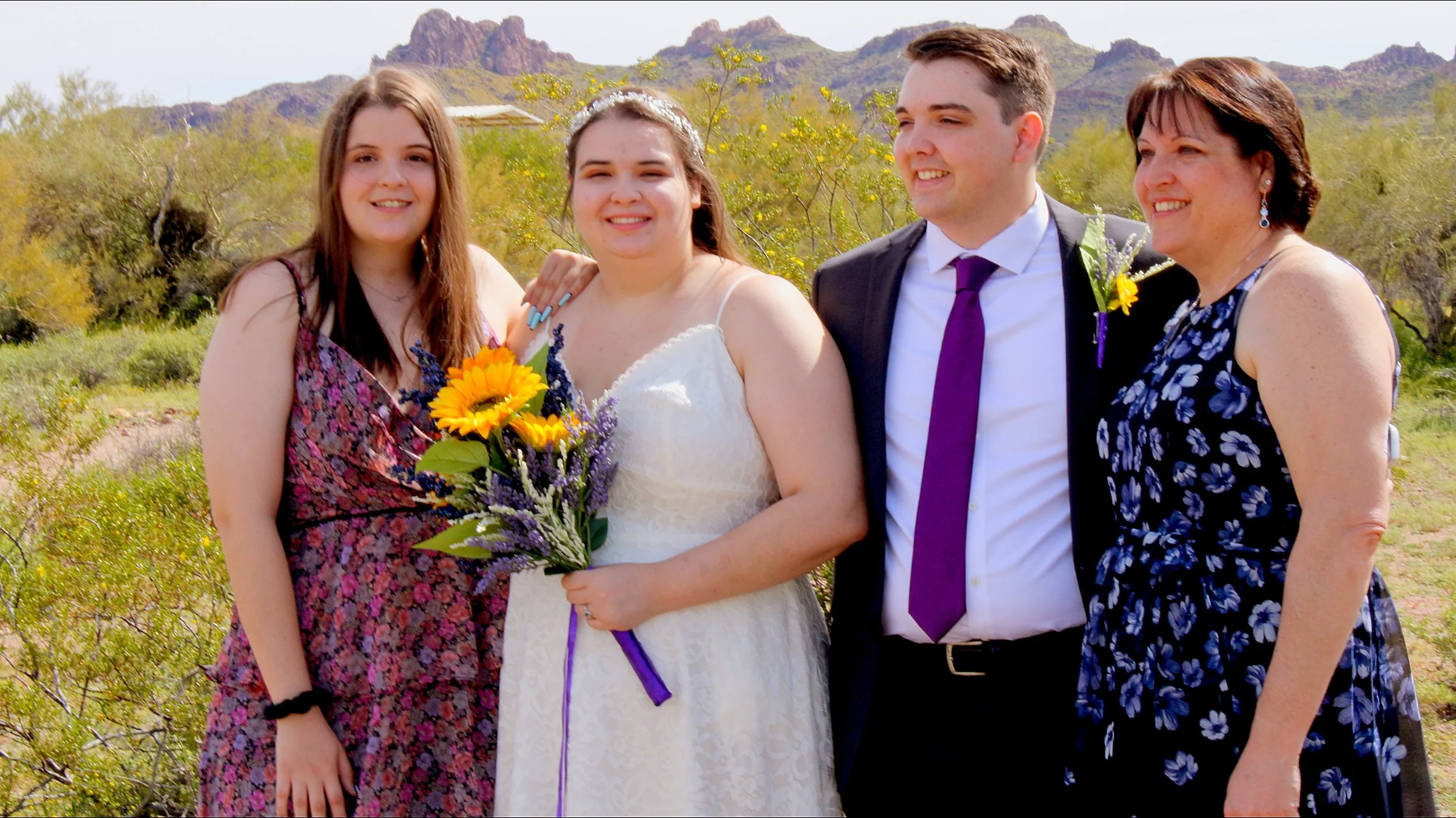 A group of four people standing outdoors in a desert landscape, with mountains in the background. Two women, one in a floral dress and one in a black floral dress, and two young adults, a woman in a white wedding dress holding a sunflower bouquet and