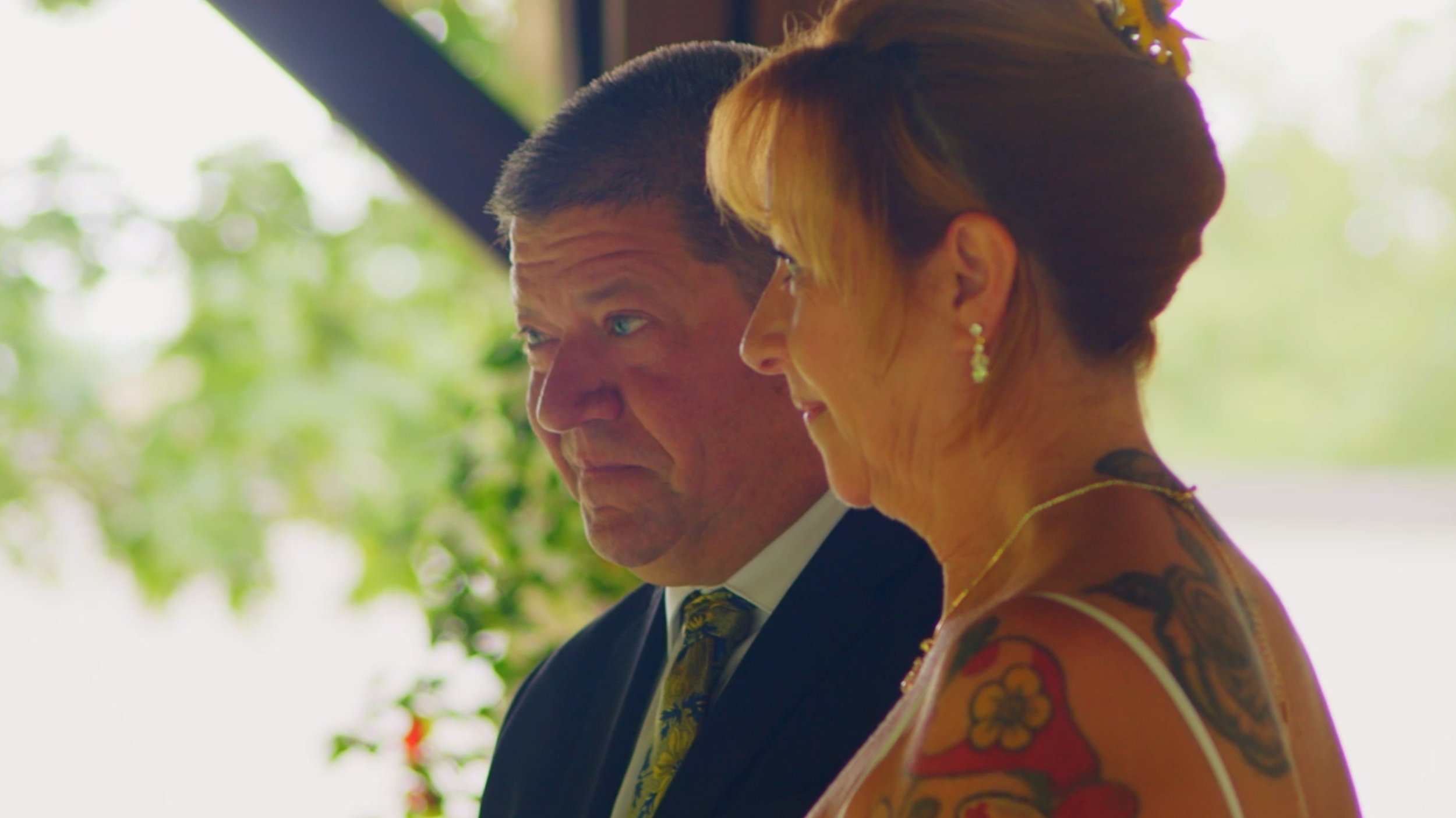 A man in a suit and a woman with tattoos and jewelry standing side by side outdoors on a sunny day.