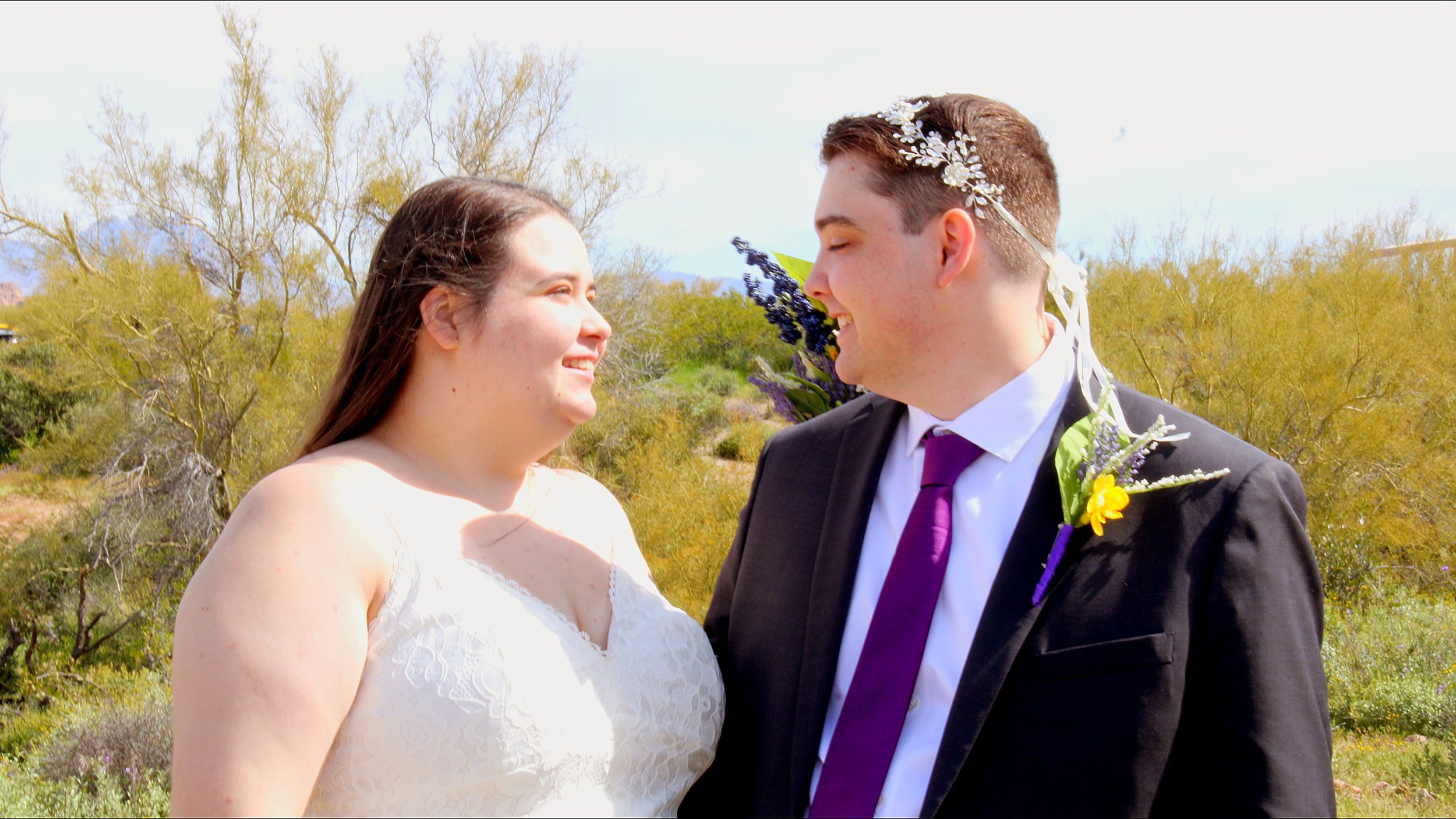 A bride and groom smiling at each other outdoors on a sunny day, with trees and mountains in the background.