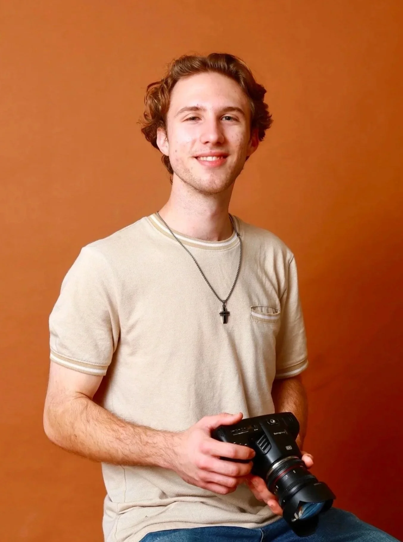 A young man with curly red hair, wearing a beige t-shirt and a cross necklace, holding a camera, standing against a plain orange background.
