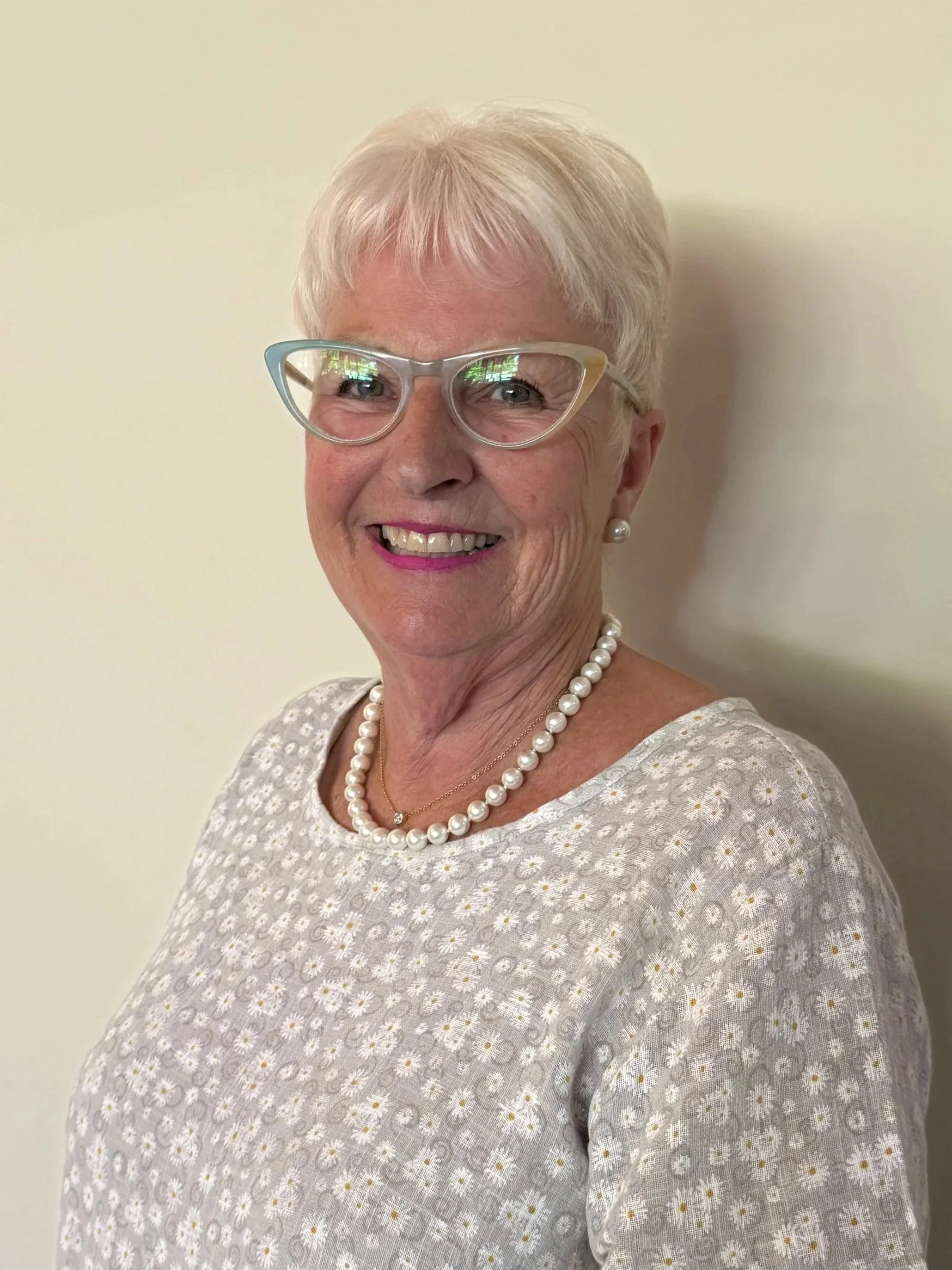 An elderly woman with short white hair, glasses, pearl earrings, and a pearl necklace, smiling in front of a plain light-colored wall.