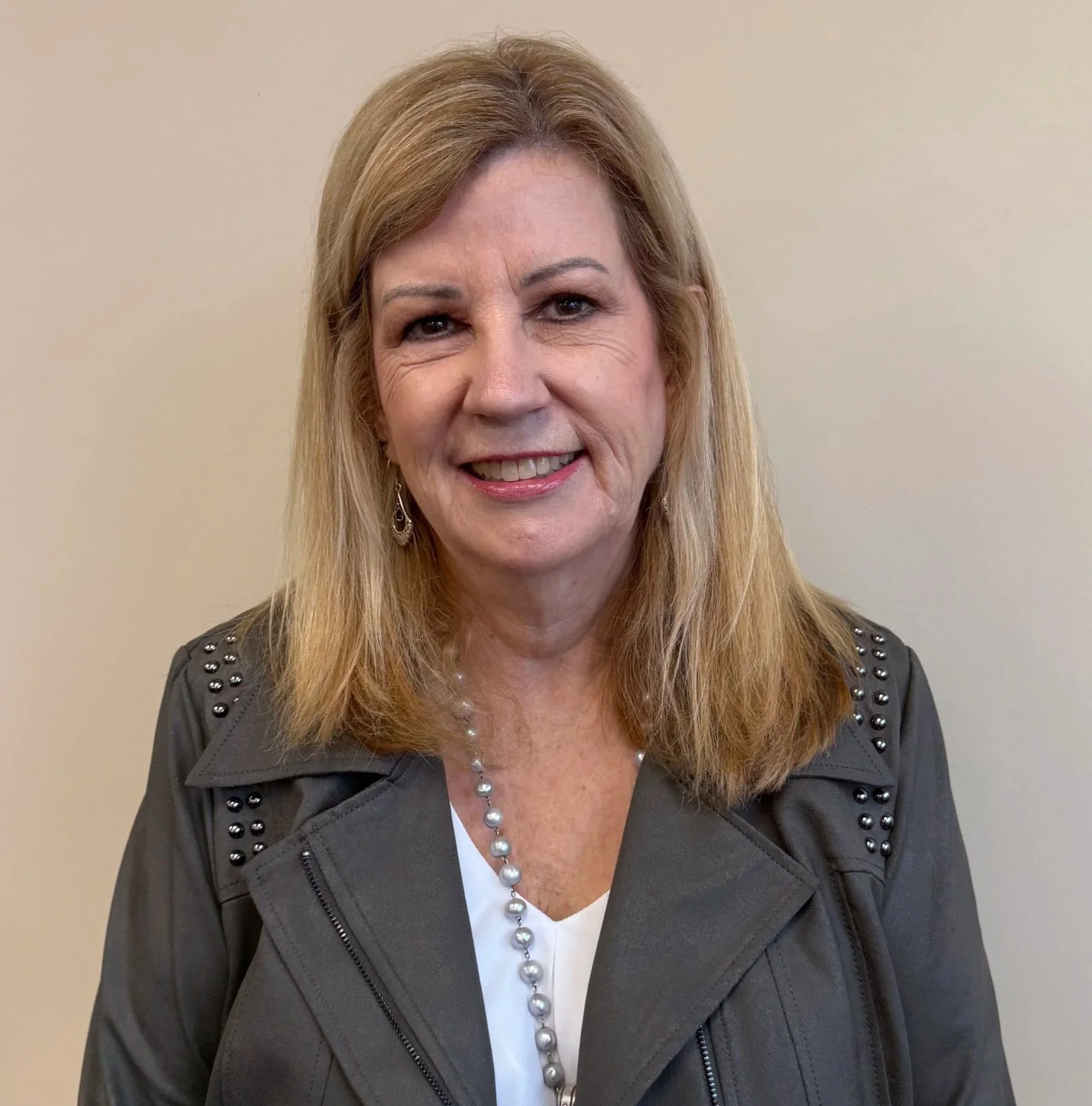 A middle-aged woman with blonde hair, smiling, wearing a grey jacket with studded detail on the shoulders, a white blouse, and a pearl necklace, standing against a plain beige wall.