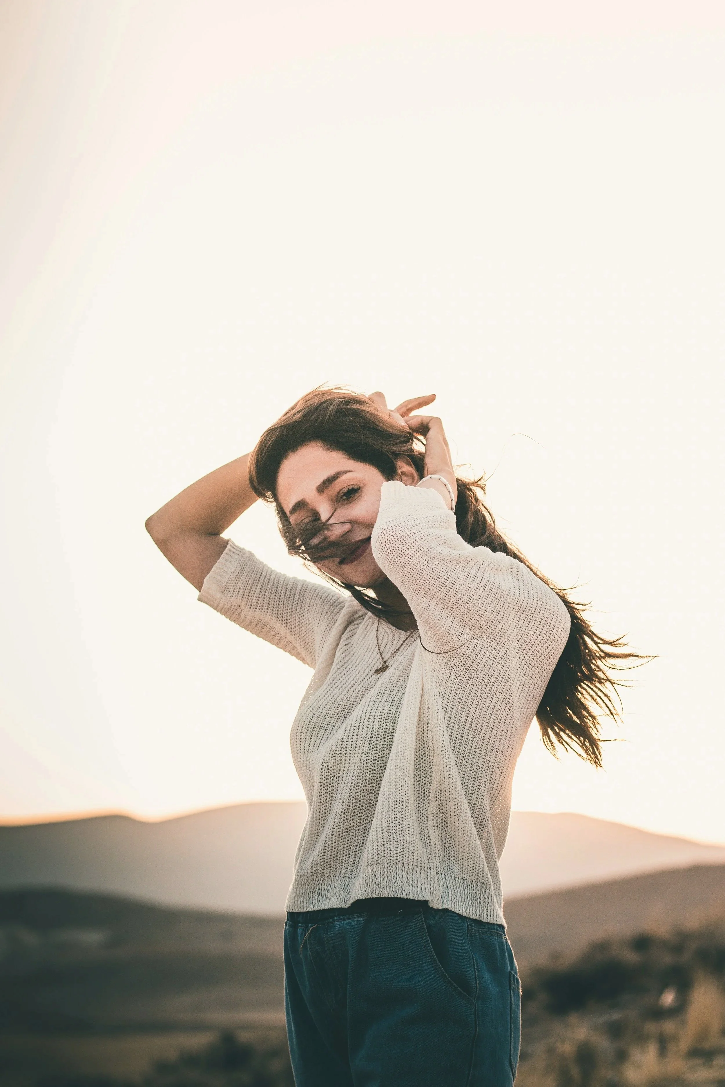 A woman with dark hair and dark lipstick smiling outdoors during sunset, wearing a white knit sweater and jeans, standing against a mountainous landscape.