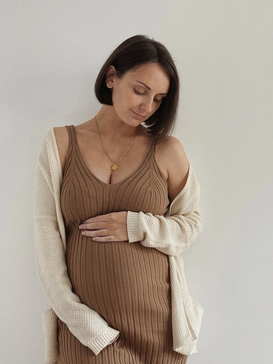 A pregnant woman wearing a ribbed brown dress and a cream-colored cardigan, standing against a plain white wall, gently resting her hand on her belly, with a serene expression.