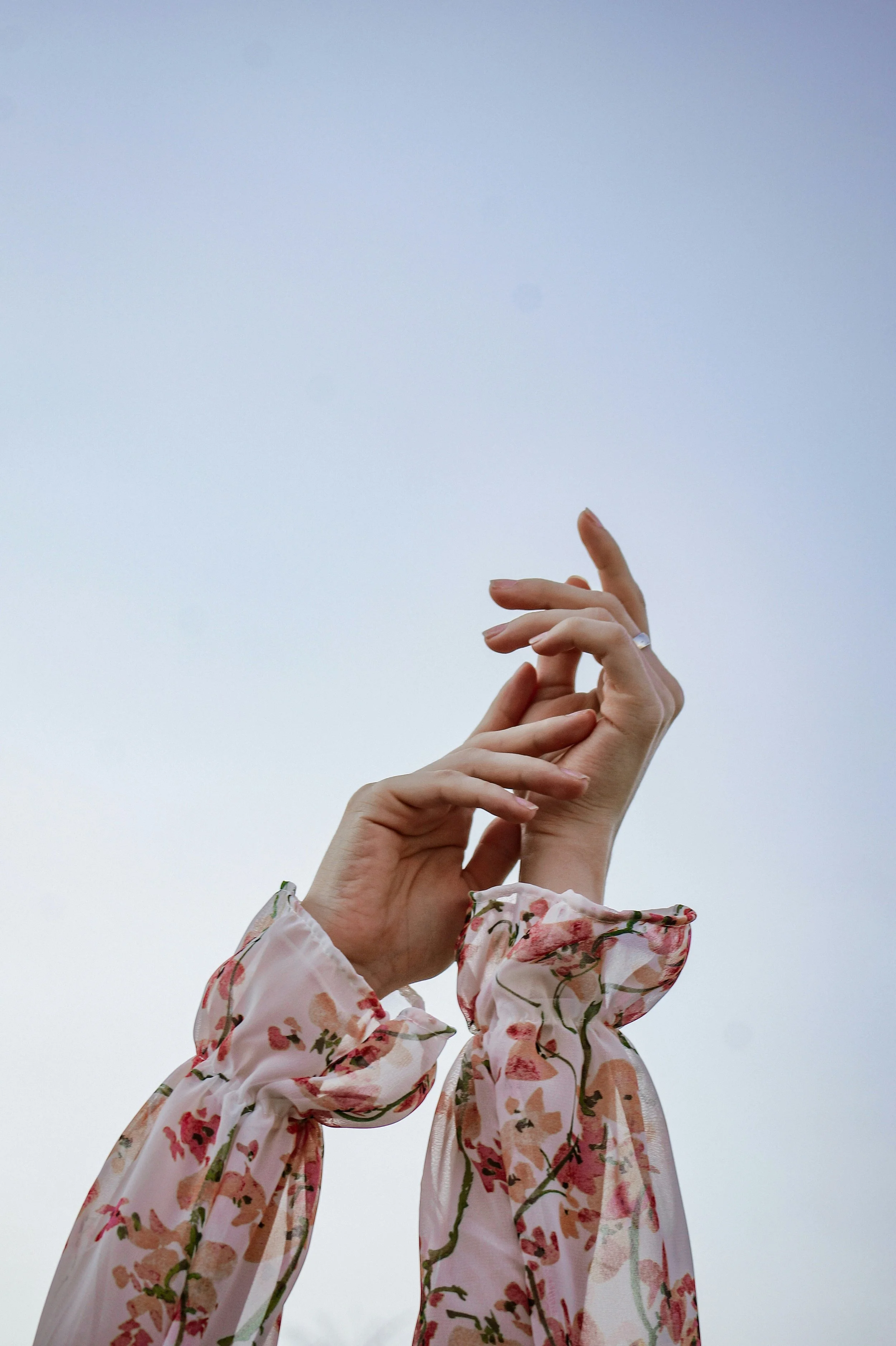 Close-up of two hands raised against a pale blue sky, dressed in floral-patterned, long-sleeved, transparent fabric.