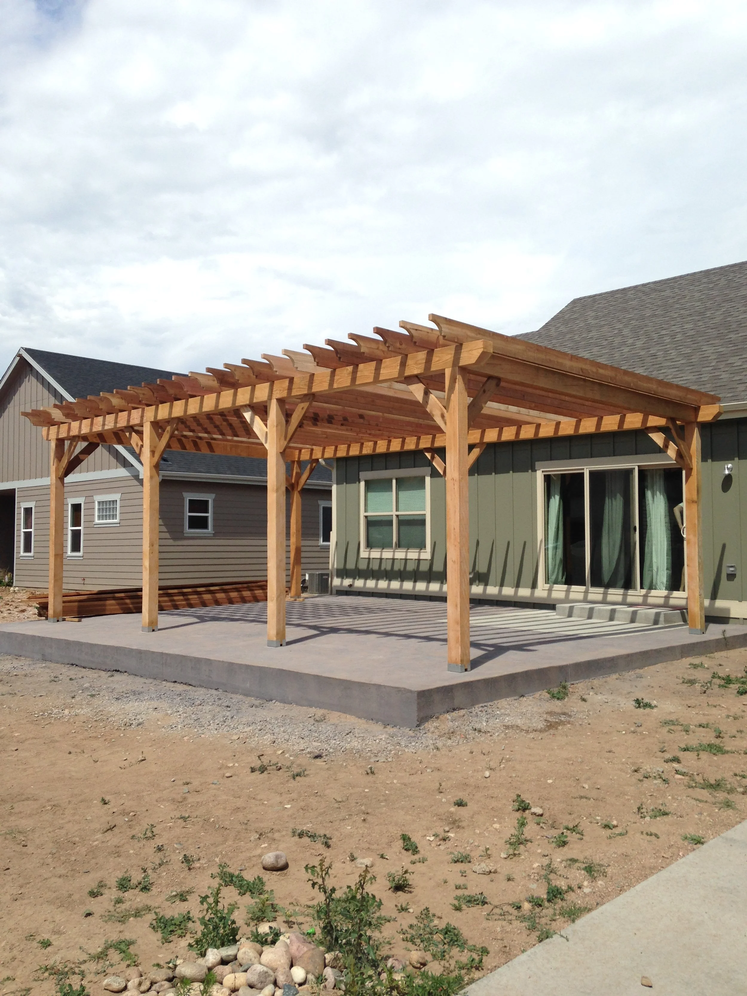 Wooden pergola structure built over a concrete patio in a backyard, with neighboring houses visible and a partly cloudy sky overhead.