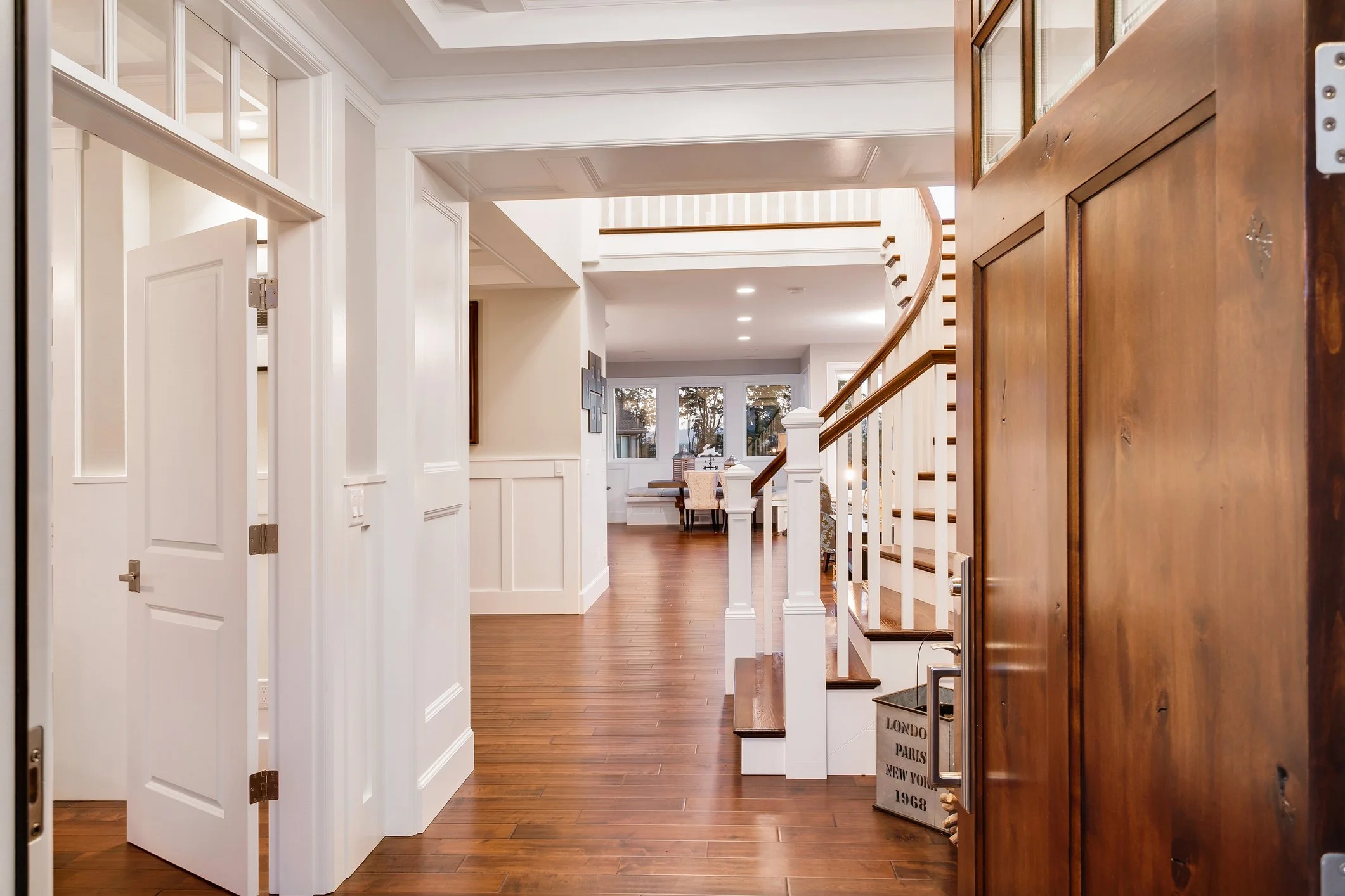 Entrance hall with hardwood flooring, white walls, and a staircase leading to an upper level, with a view into a dining area with large windows. Main Floor Whole Home Renovation