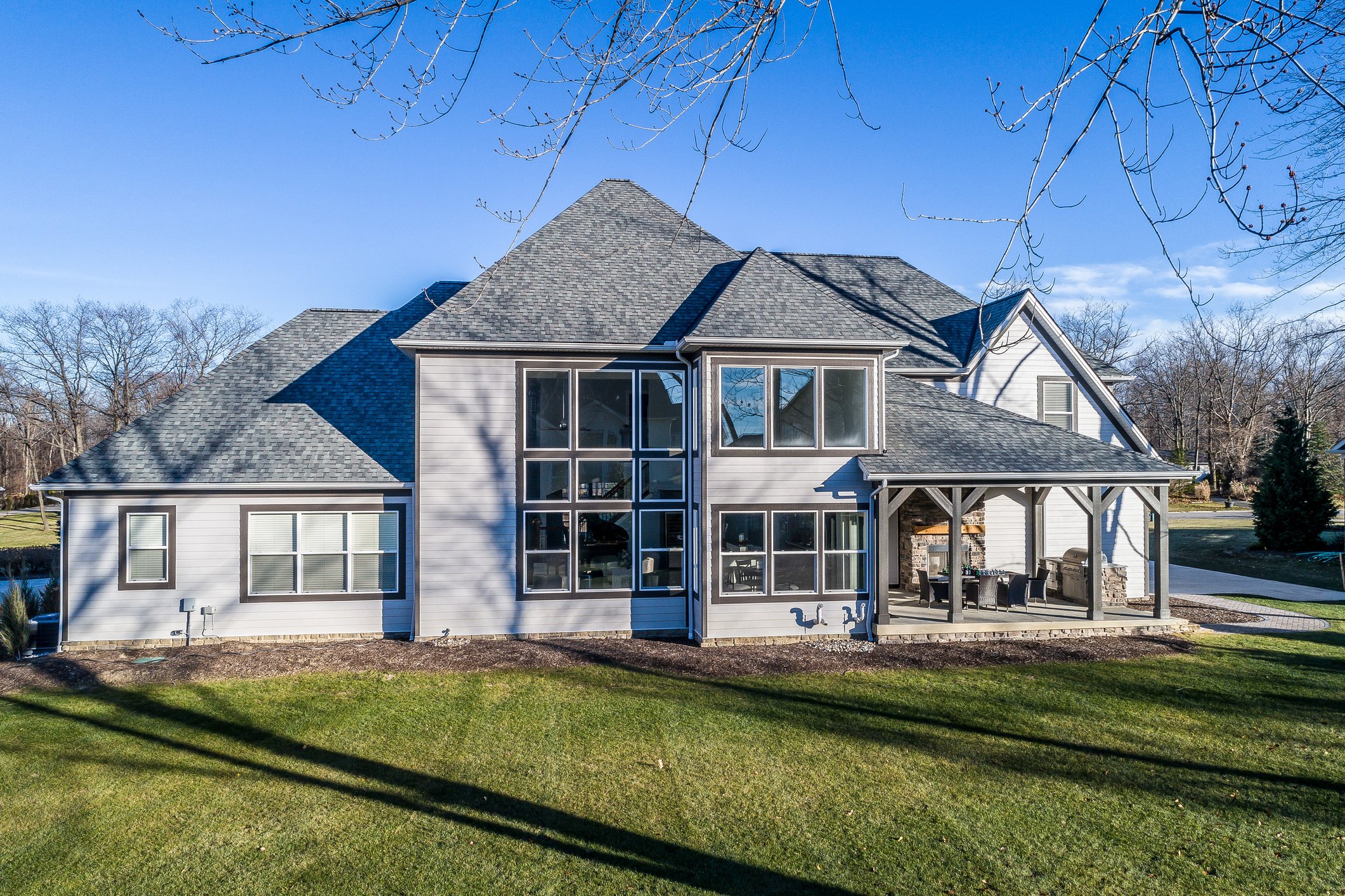 Modern two-story home addition with gray exterior, large windows, and a covered patio with outdoor furniture, set in a green yard with trees.