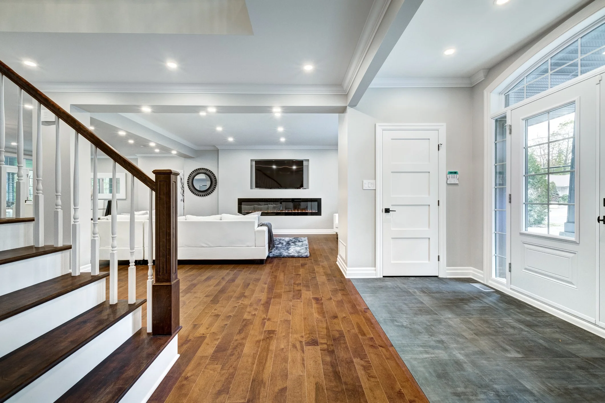 Modern living room with hardwood flooring, white walls, a staircase with wooden steps, a white sectional sofa, a fireplace, a wall-mounted TV, and large windows with white frames. Whole Home Renovation in Ontario