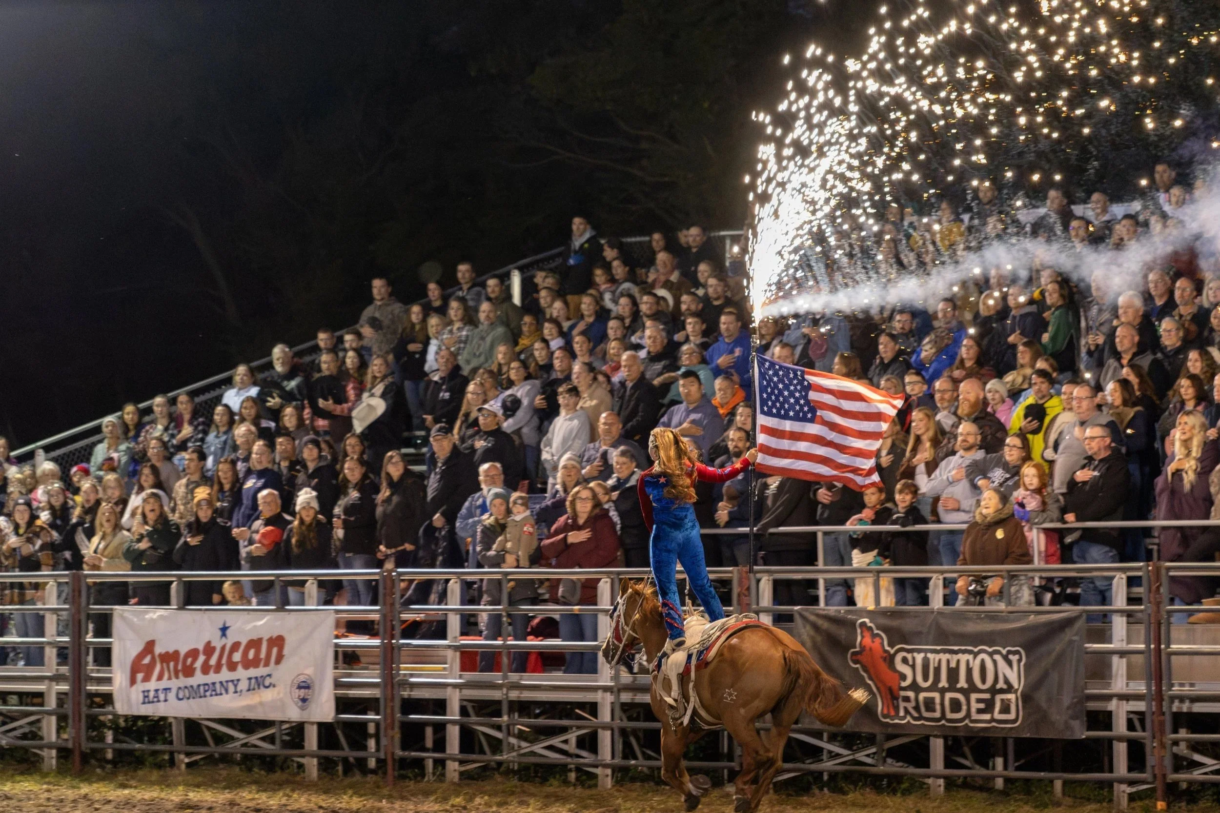 A rodeo event at night with a large crowd watching. A woman dressed as Wonder Woman is riding a bucking horse in the center, holding an American flag. Sparks are flying from a firework display nearby.