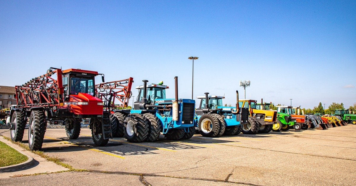 Rows of tractors and farm equipment lined up across a paved auction lot under a clear blue sky with wide open space.