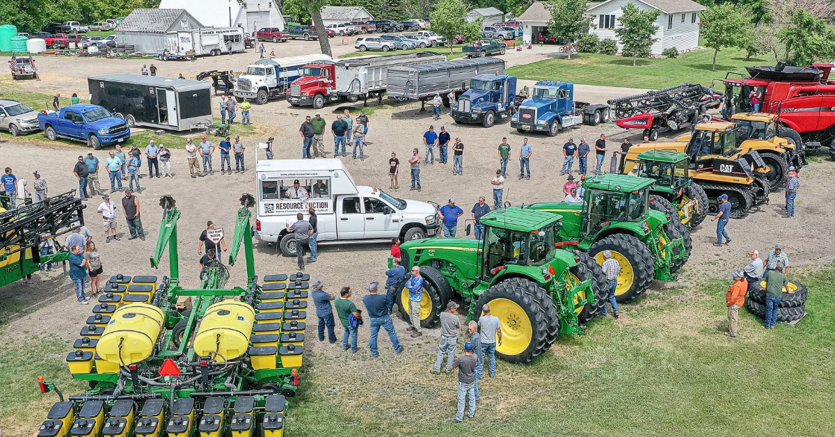 Large crowd gathered at a farm equipment auction featuring tractors, planters, construction equipment, and active bidding.
