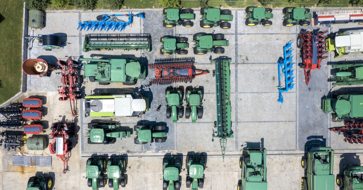 An overhead view of many agricultural vehicles. Many of them are green, and they're all sitting in a large parking lot.