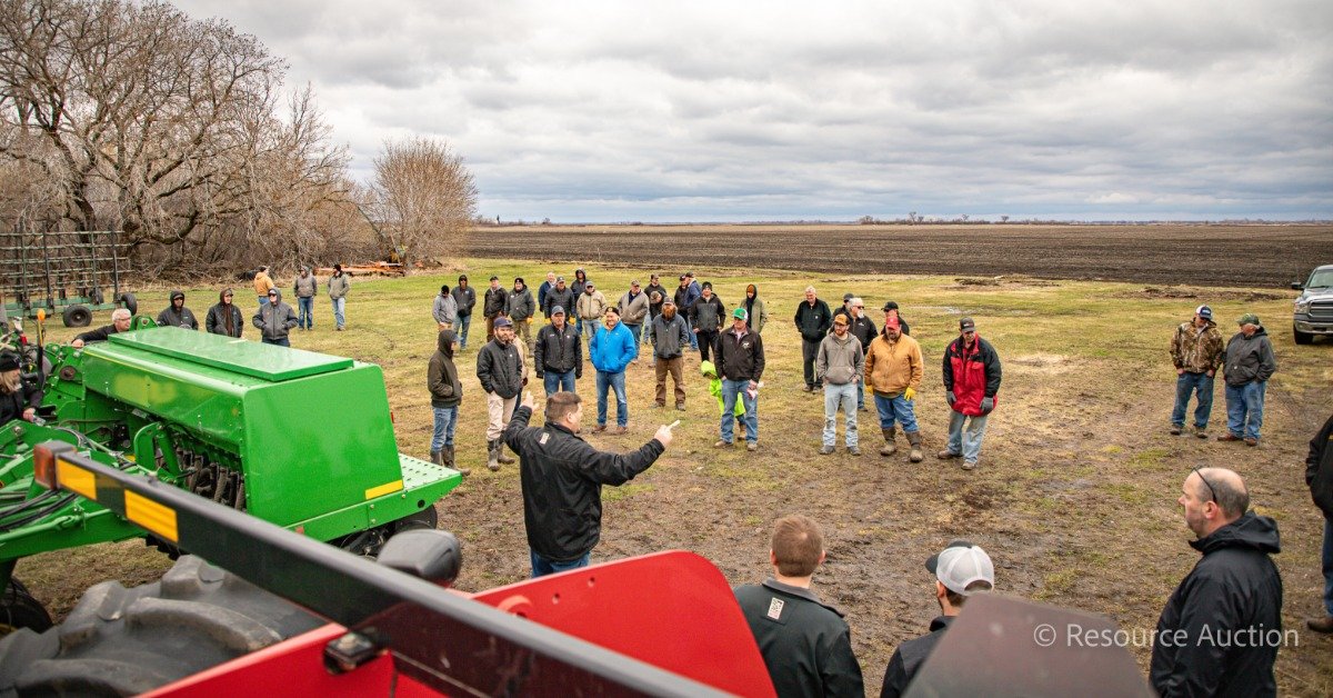 A group of people are standing in a grassy area. There is a tractor in the foreground and a large dirt field in the background.