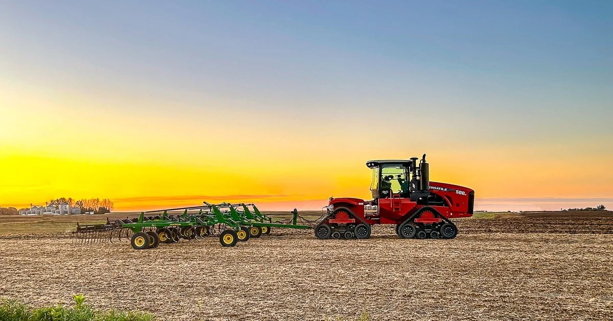 A red tracked tractor pulling a green tillage implement across a harvested field under a colorful sunset sky.