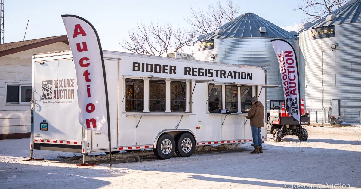 Bidder registration trailer at a farm auction site with signage, grain bins, and participants checking in before bidding.