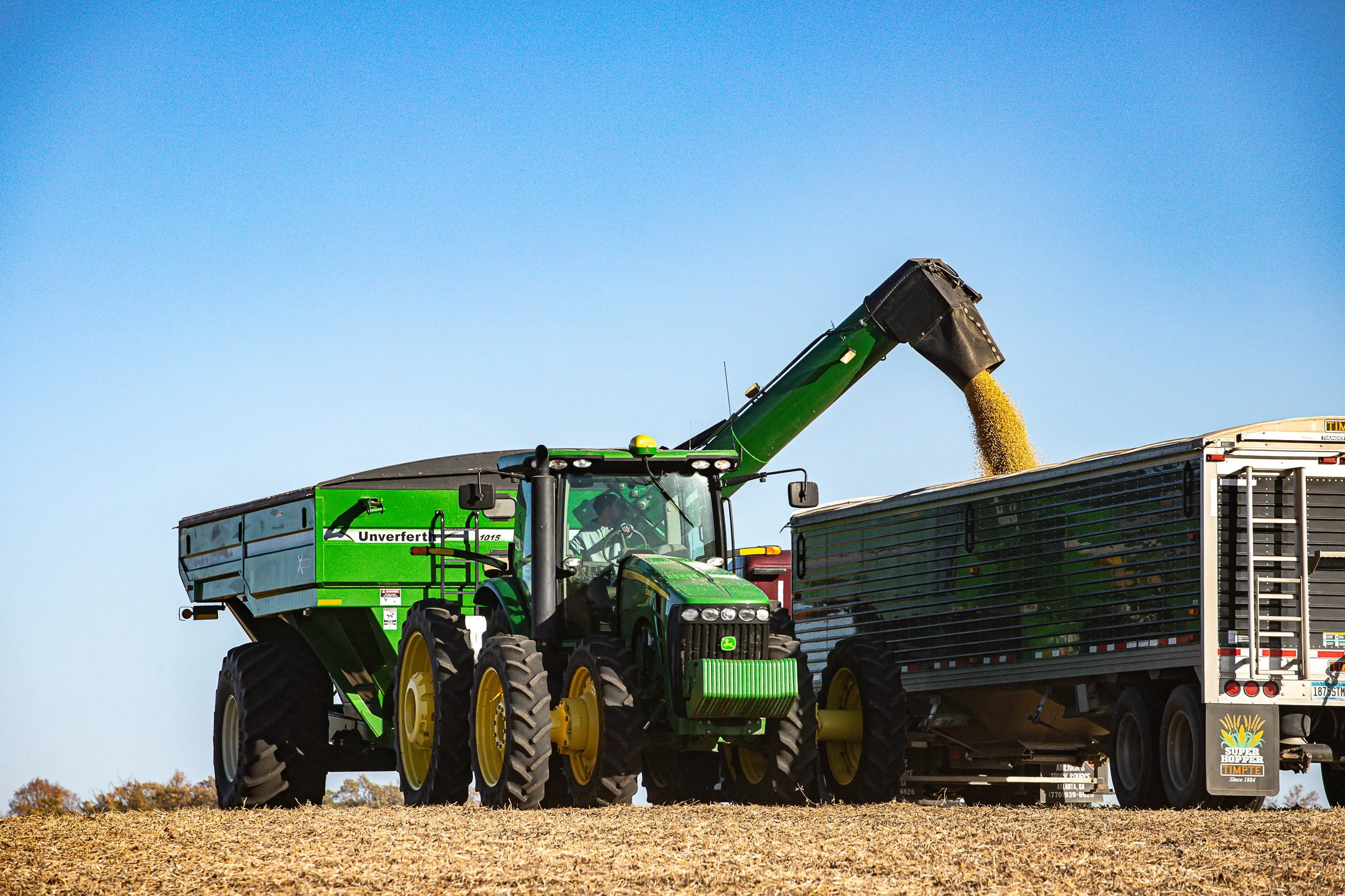A green tractor unloads grain from a raised auger into a semi trailer in the middle of a fully harvested field.