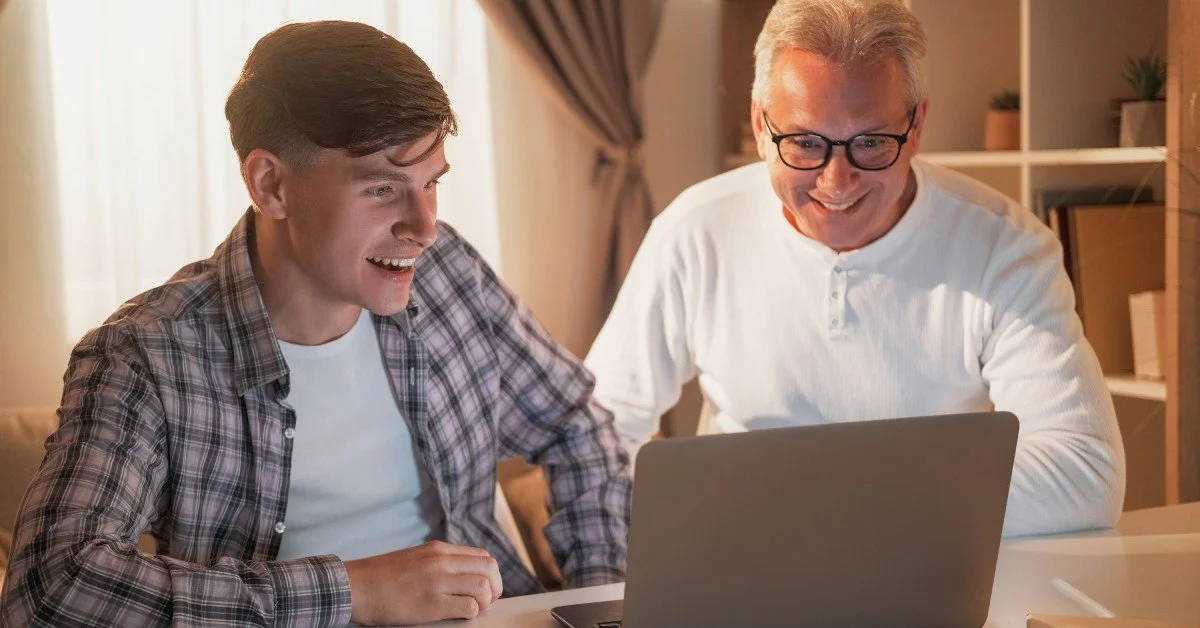 Two men sitting at a table looking at a laptop together. One is much older than the other, but both of them look excited.
