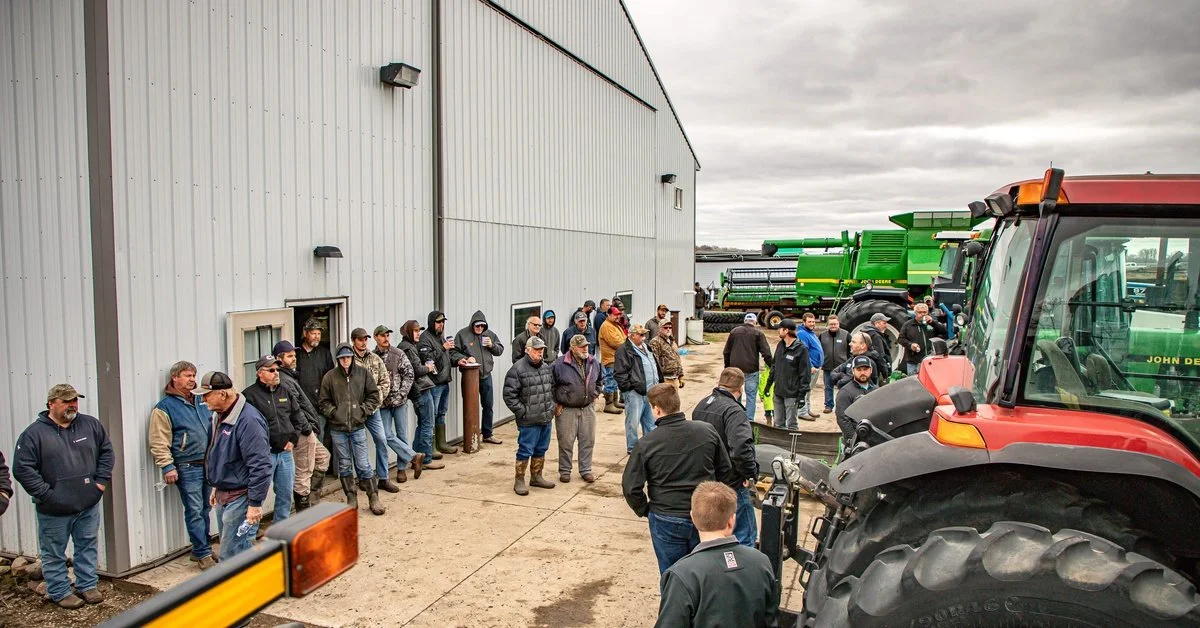 People gathering outside a metal farm building near large tractors and equipment under a cloudy sky.