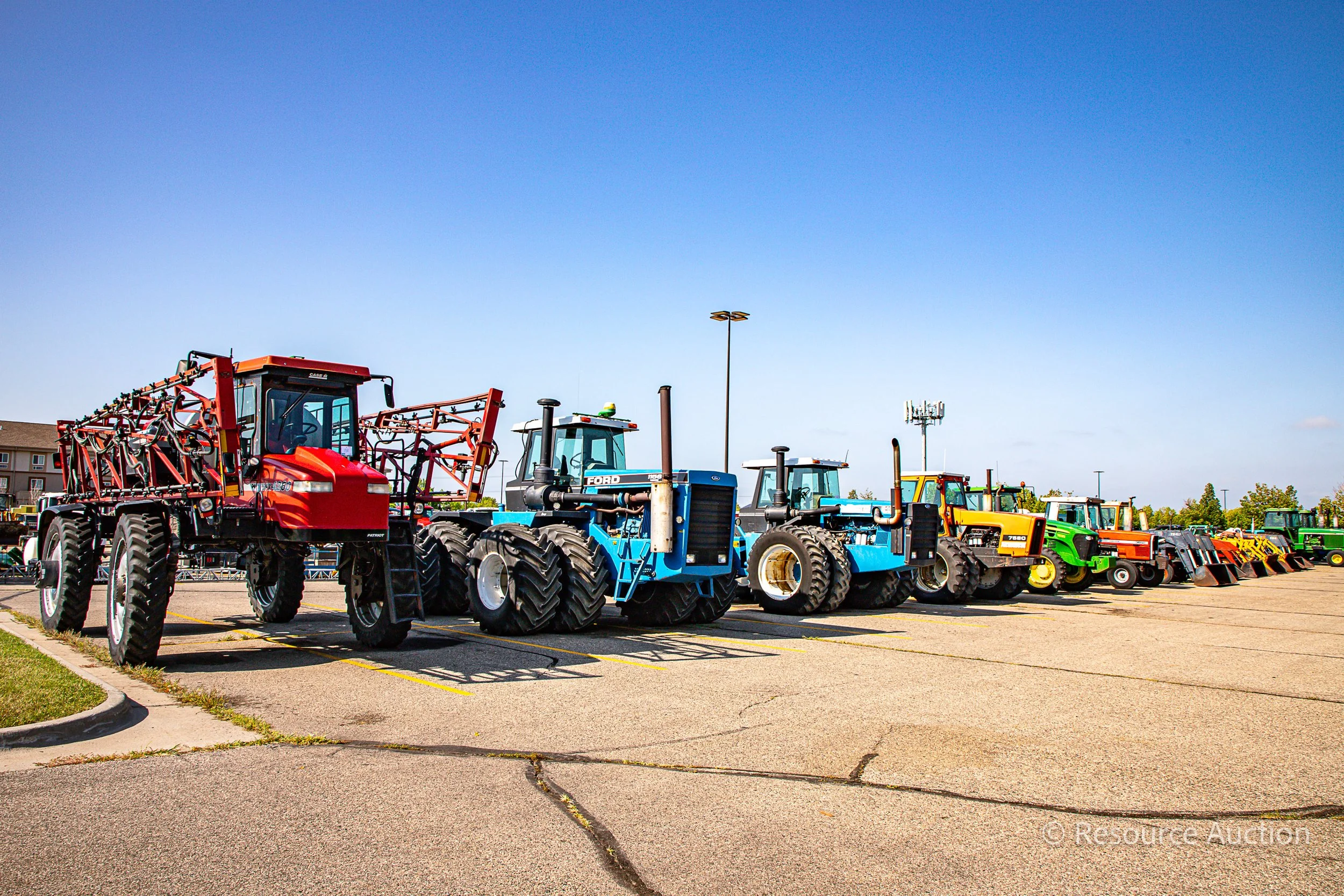 A line up of different colored farming vehicles parked next to each other. They are situated in place for an auction.