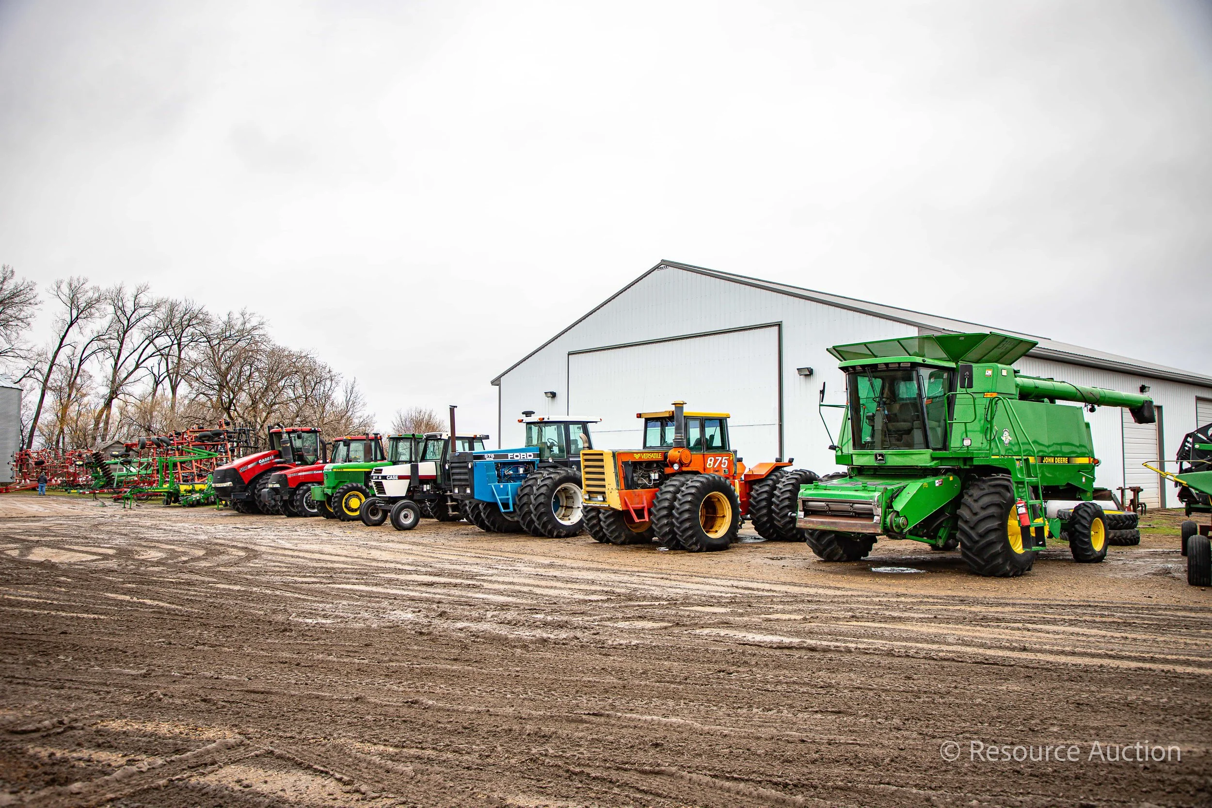 Farm equipment lines a muddy lot outside a large white building, containing a variety of tractors and combines.