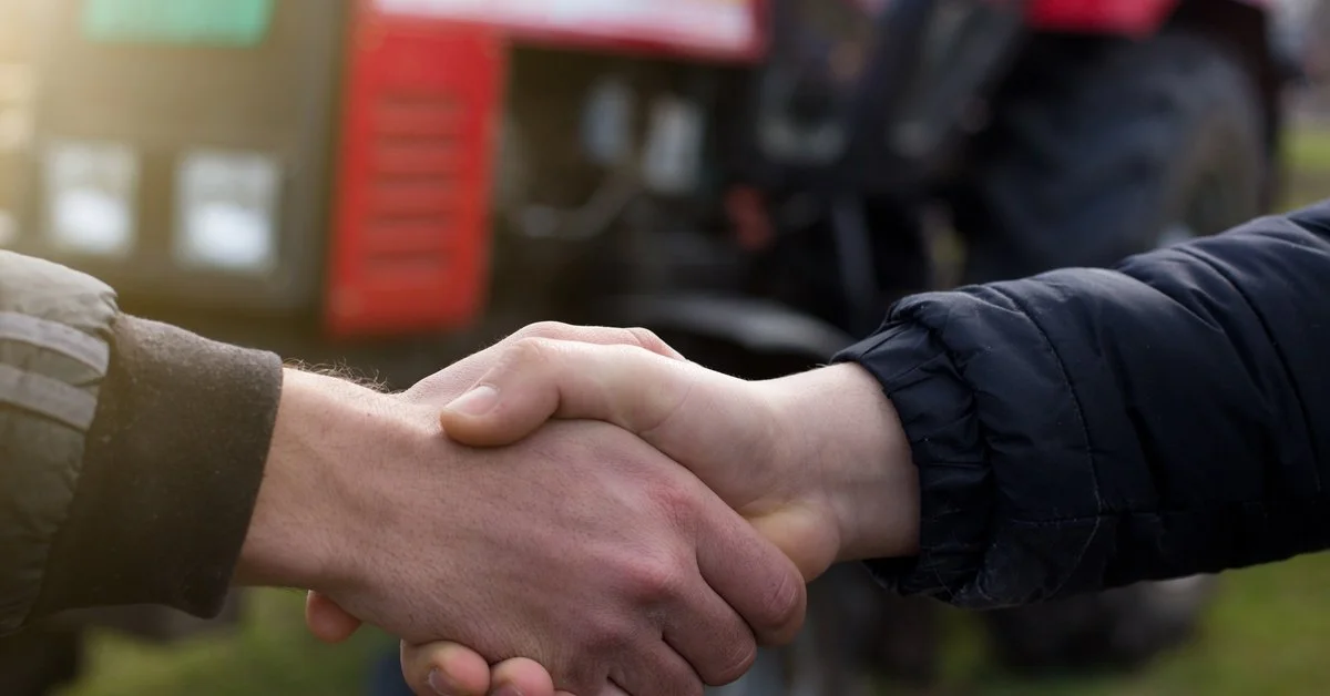 A close-up of two farmers shaking hands. There is a red tractor behind them, but it's blurry and hard to see.