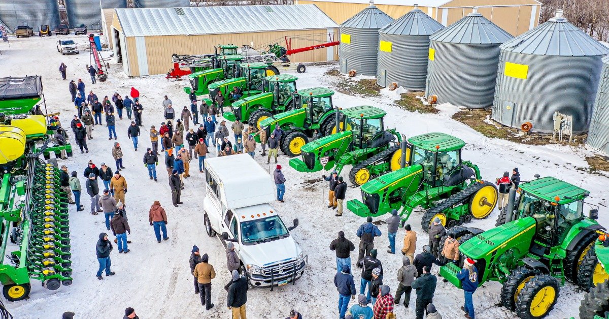 People walking among green tractors and farm equipment in a snowy farmyard with metal grain bins and buildings nearby.