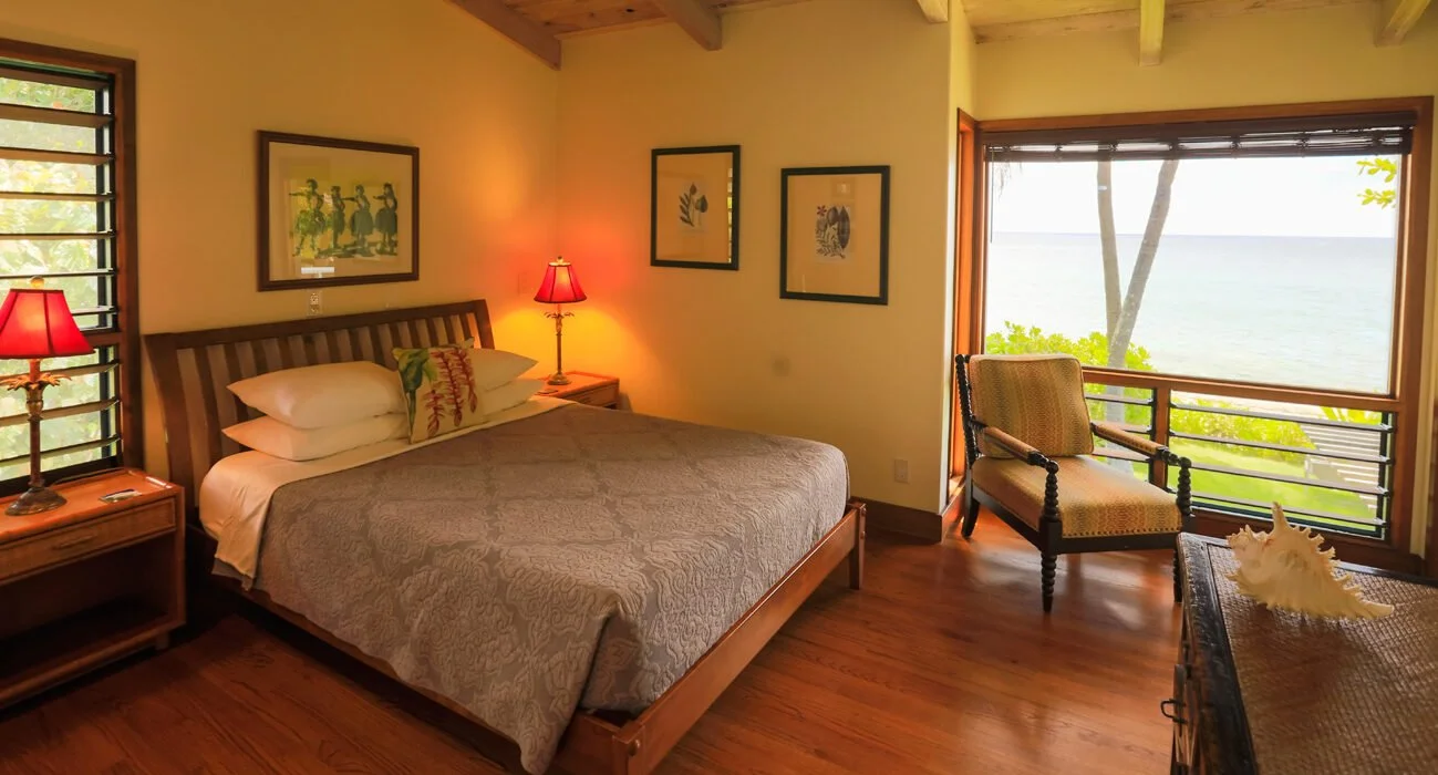 Hotel room with a large bed, framed artwork, two bedside lamps, a sitting chair by a large window overlooking the ocean, and wooden furniture.