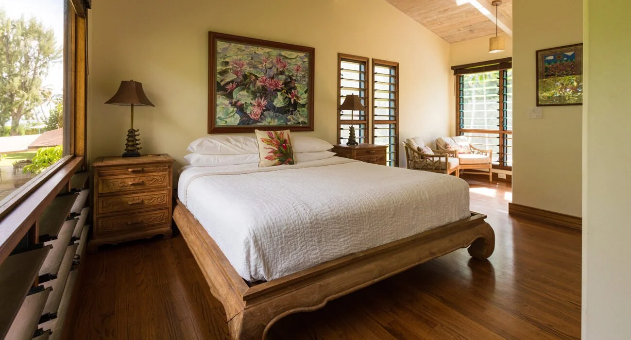A bedroom with a wooden bed, white bedding, colorful pillows, and wooden furniture, including nightstands with lamps. There are windows with wooden blinds and a sitting area with chairs and a small table by a large window.