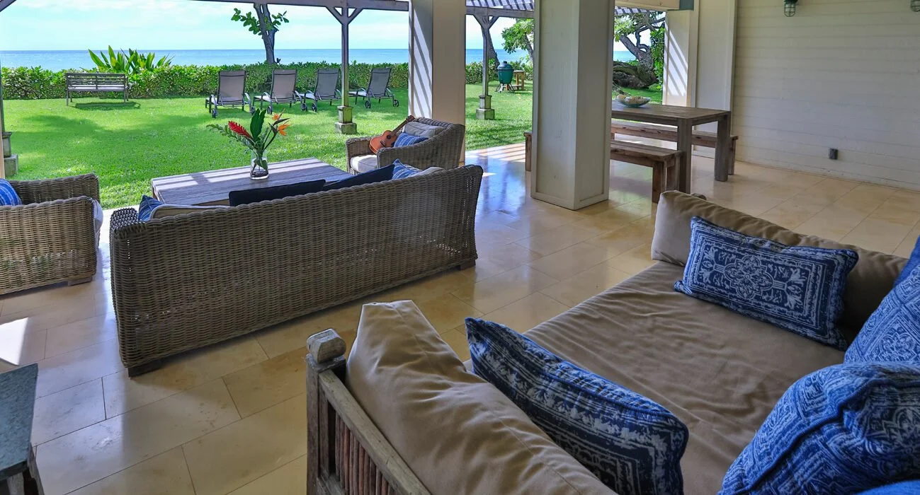 Living room with wicker and upholstered furniture, overlooking a grassy yard with lounge chairs, trees, and ocean in the background.