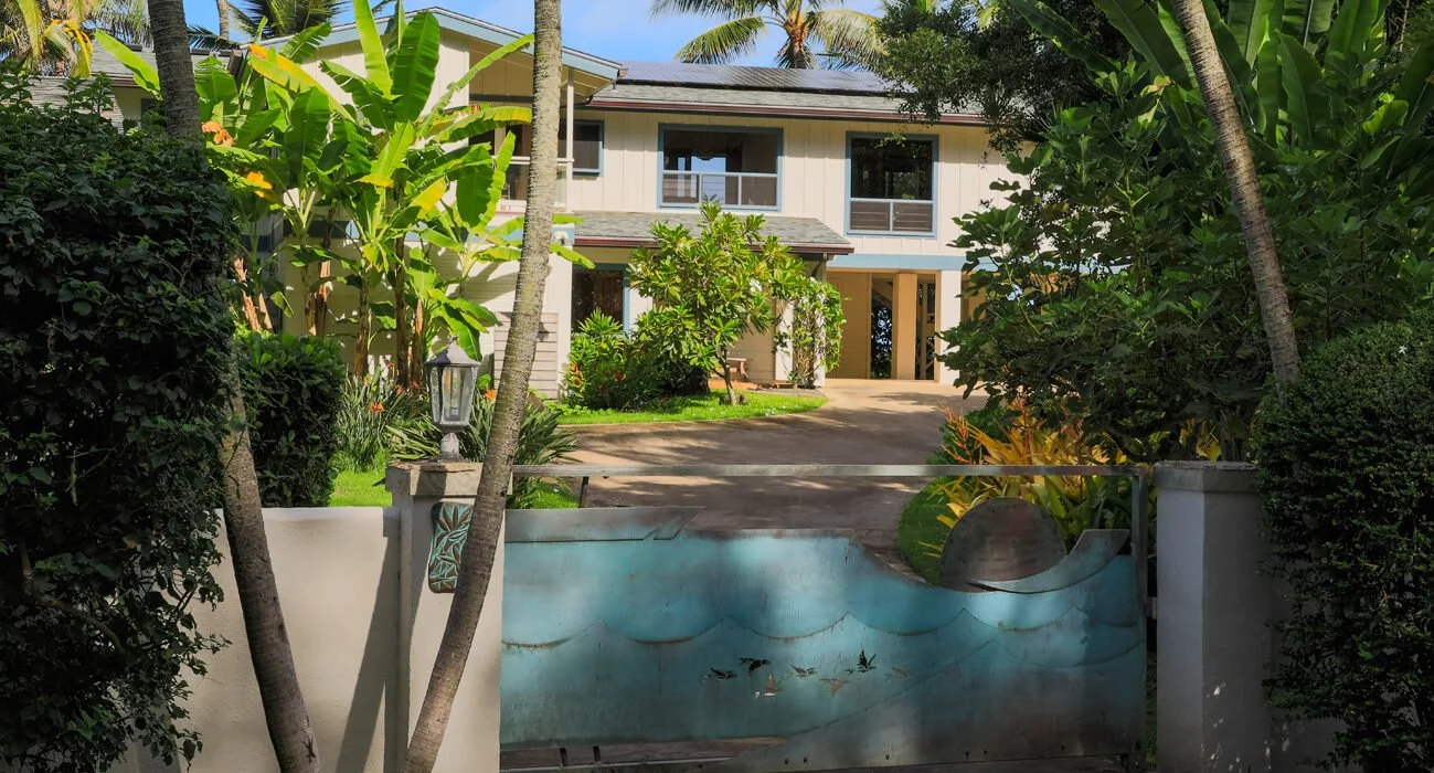 View of a two-story house surrounded by tropical plants and trees, with a decorative metal gate featuring ocean waves in the foreground.