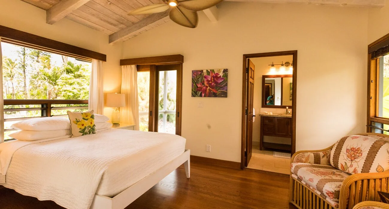 A cozy bedroom with a large bed, patterned pillows, and a nightstand with a lamp. The room has wooden flooring, a ceiling fan, and large windows revealing greenery outside. There is a floral painting on the wall and an open door leading to a bathroom
