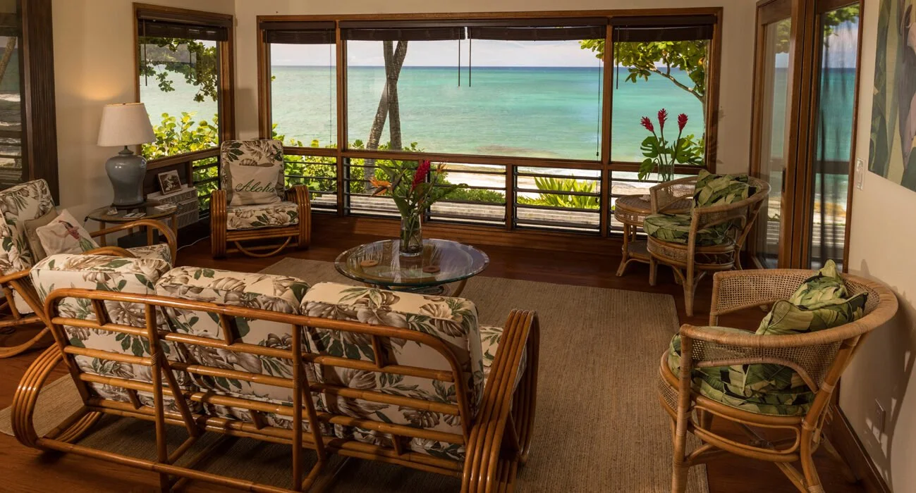 Living room with rattan furniture and tropical-themed cushions, large windows overlooking a beach with trees and ocean, and a glass coffee table with a flower arrangement.