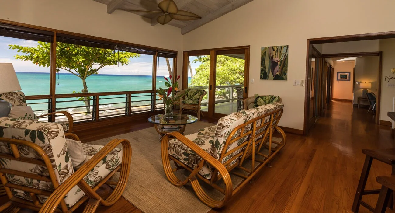 Living room with large windows overlooking a tropical beach with turquoise water, a tree, and a clear sky, featuring tropical-patterned furniture, a glass coffee table with a flower arrangement, and wooden floors.