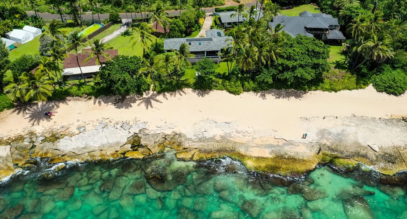  aerial view of a tropical beach with white sand, clear turquoise water with rocks, and lush greenery with palm trees and houses in the background