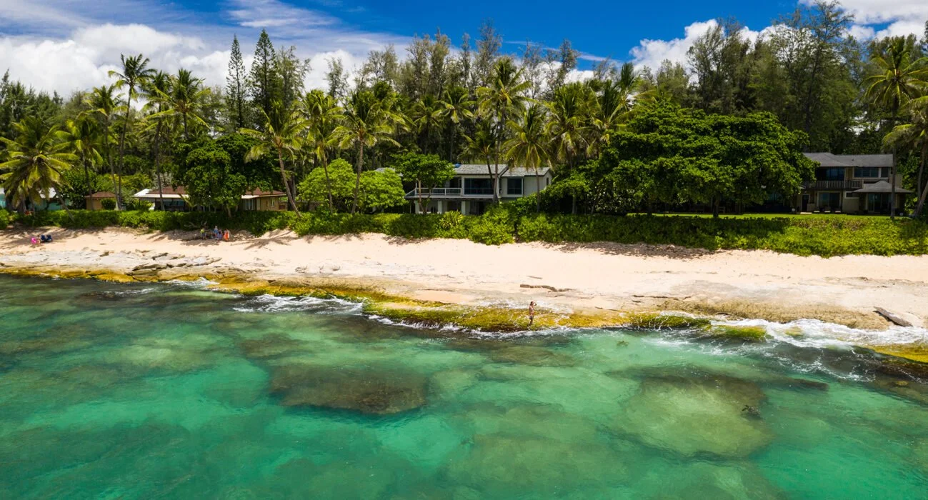 Tropical beach with white sand, clear green water, lush greenery, and houses among palm trees under a partly cloudy sky.