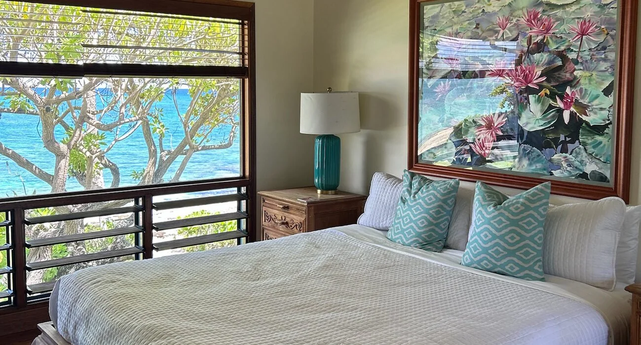 A bedroom with a large window showing a view of a tree and water outside. The bed has white linens and three decorative pillows with a blue and white pattern. A wooden nightstand with a teal lamp is beside the bed. A framed painting of pink and purpl
