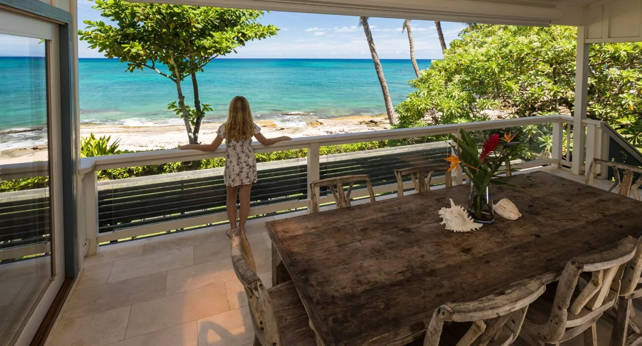 A young girl stands on a balcony, looking out over a beach with turquoise water and white sand, surrounded by green trees, with a rustic wooden table and chairs inside.