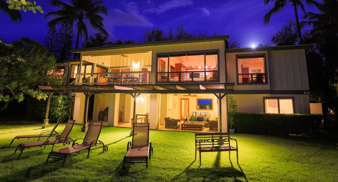 A modern two-story house at night with large glass windows, outdoor lounge furniture, and lawn chairs on a well-lit grassy yard, surrounded by palm trees.
