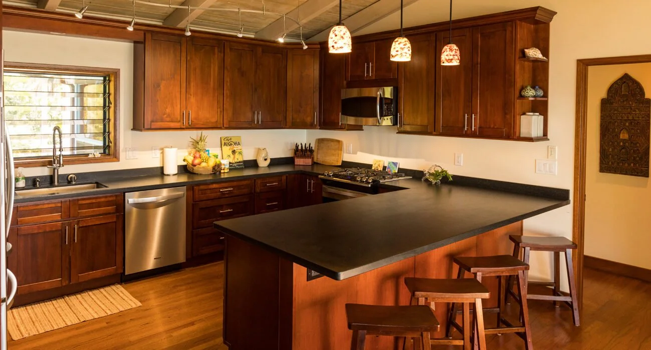 Kitchen with wooden cabinets, black countertop, stainless steel dishwasher, microwave, and sink. There are three hanging pendant lights with decorative shades, a window with louvered blinds, and three barstools at a breakfast bar. Decor includes a pi