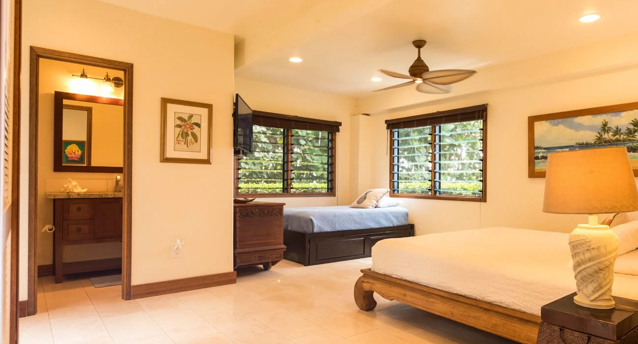 A bright hotel room with a large bed, bedside lamp, windows with wooden blinds, a ceiling fan, and a smaller bed against the wall.