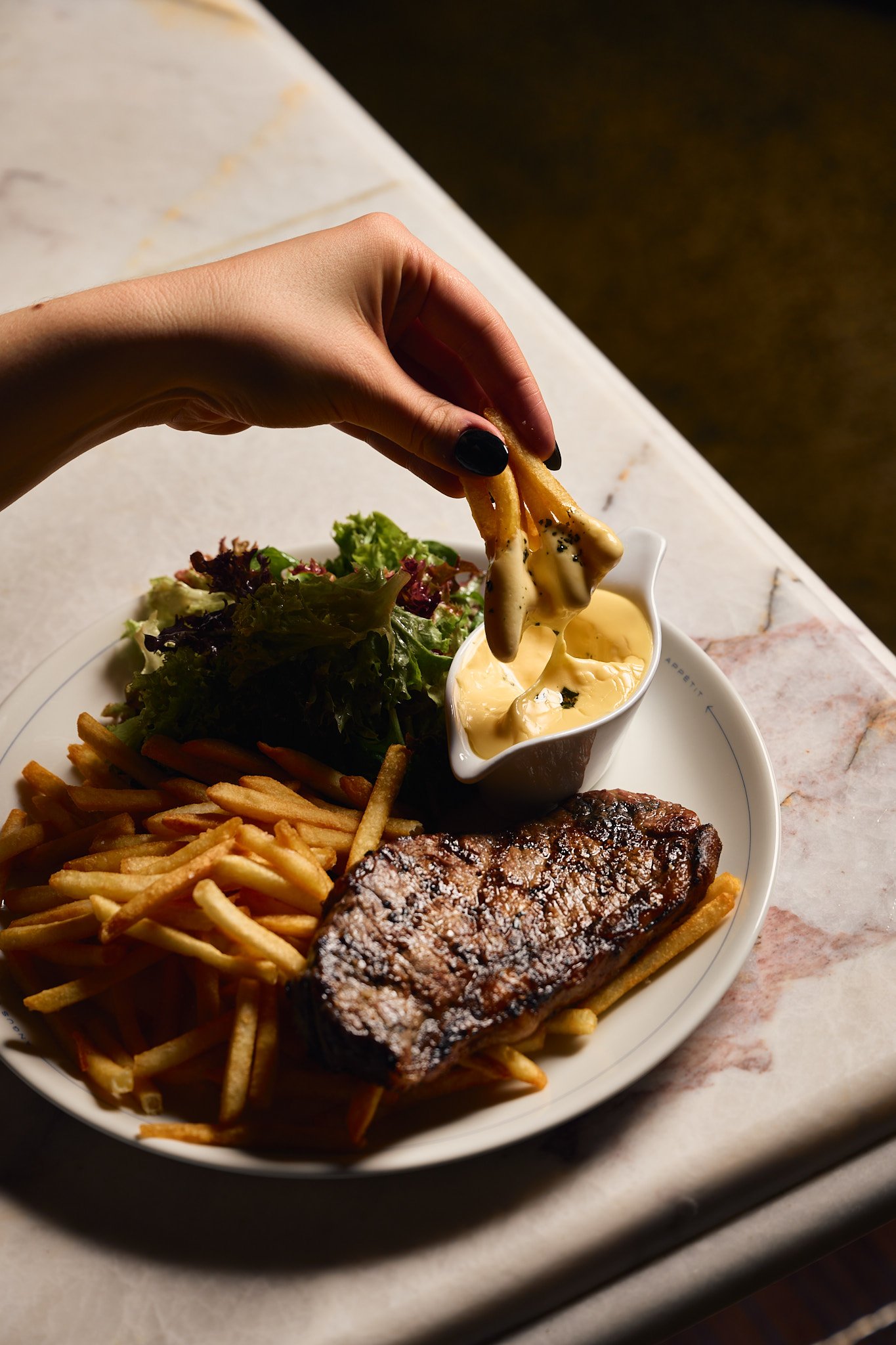 Angus & Bon Signature grilled steak, French fries, a green salad, and a small container of creamy dipping sauce, with someone dipping French fries into the sauce.