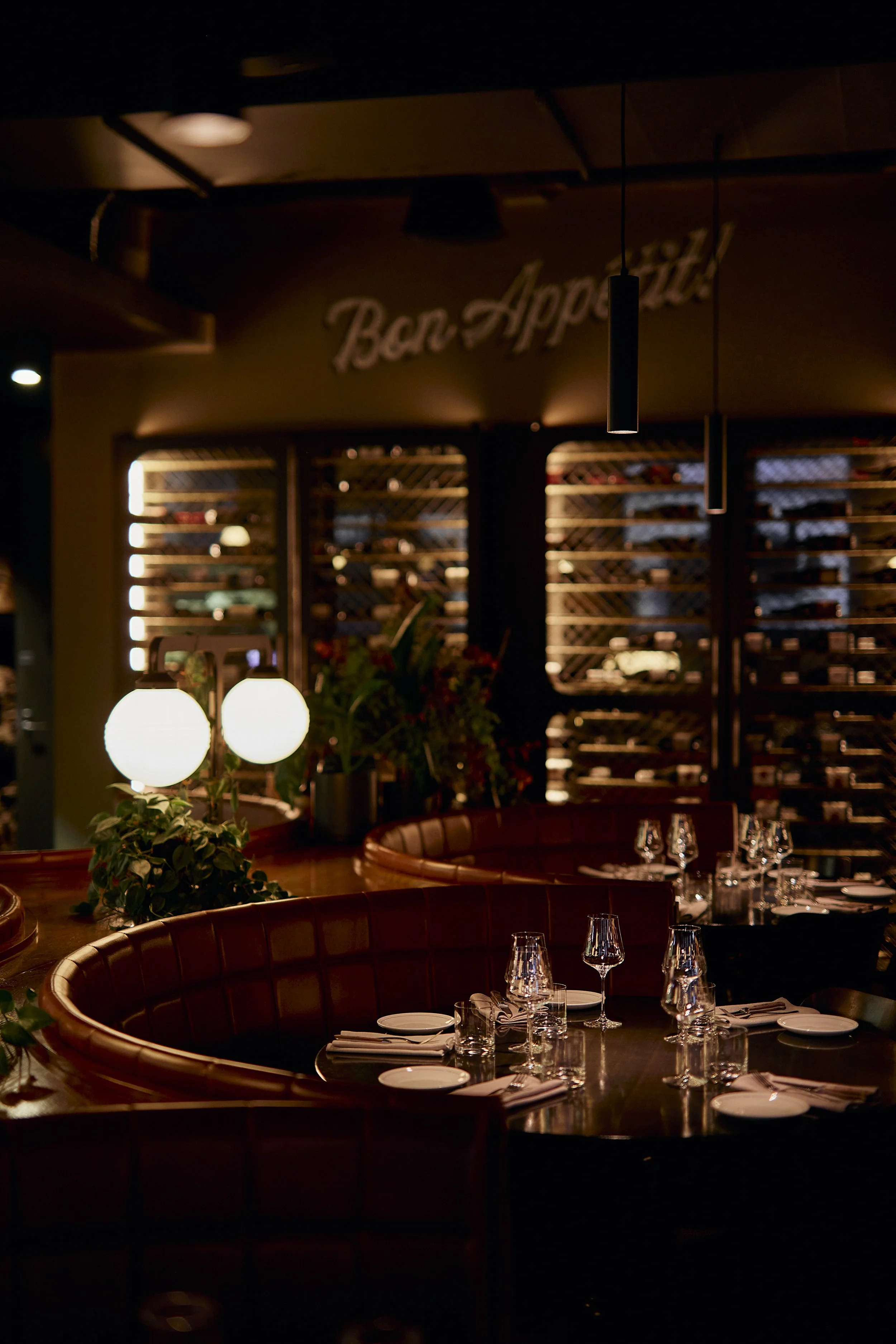 Interior of a restaurant with table settings, wine glasses, and dim lighting, featuring a curved leather booth and decorative lighting fixtures.