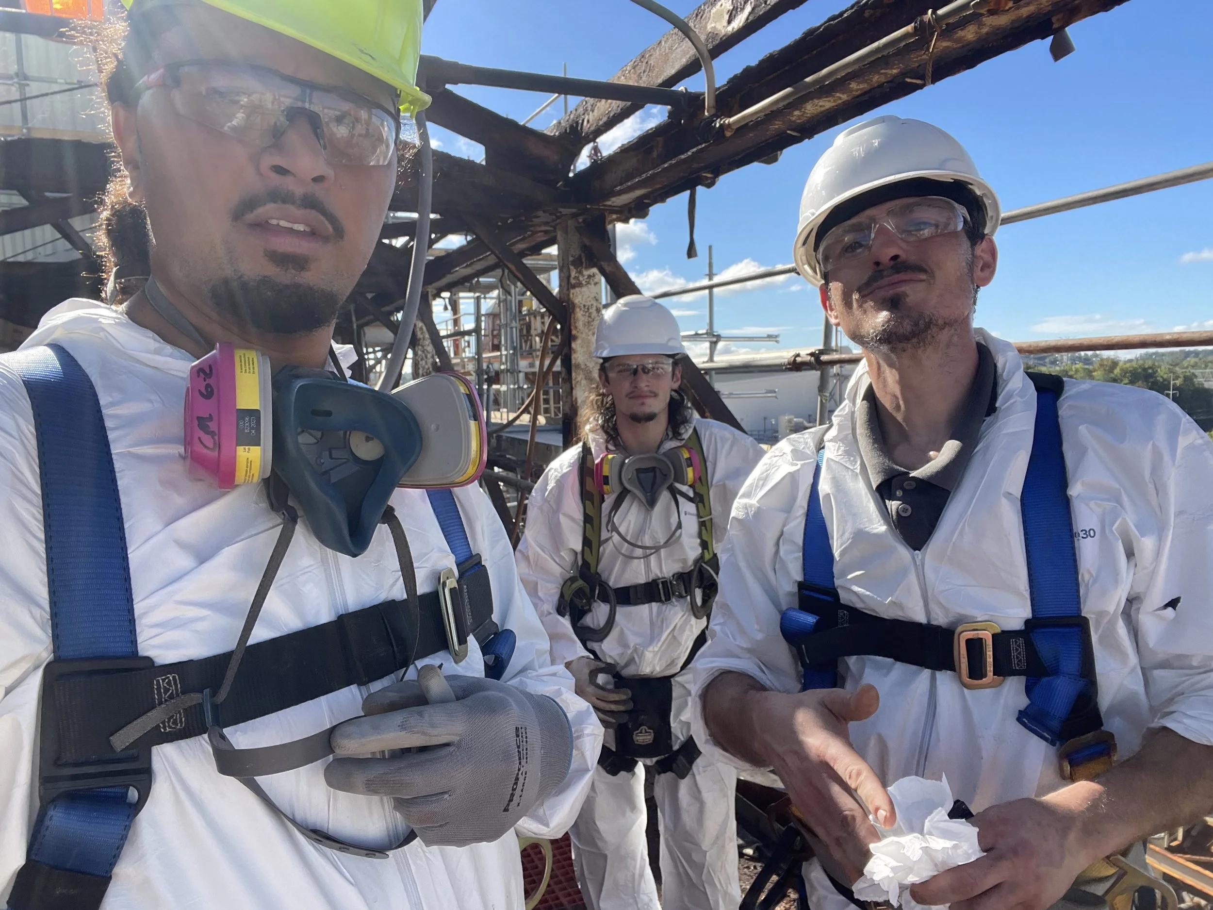 Three industrial workers in white protective suits, hard hats, safety glasses, respirators, and harnesses pose at a construction site.